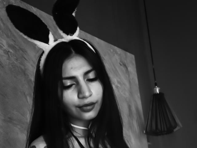 BenedettoMichelRuby with long dark hair, wearing bunny ears, closed eyes, subtle smile, and a choker, in a black-and-white photo, showing her smooth, cute, and sexy face.