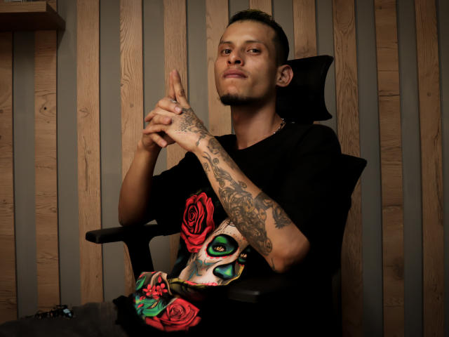 BenedettoMichelRuby with tattoos on arms, wearing black shirt, skull mask with red roses, sitting in chair, hands clasped, short dark hair, wooden wall, confident smirk.