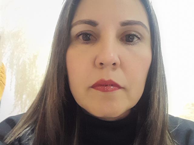 El rostro de AdelaShery, con labios rojos brillantes, ojos marrones y cabello castaño liso, lleva un cuello alto negro y mira ligeramente hacia abajo. Su nariz está centrada..
