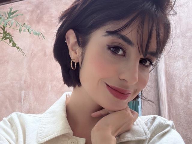 Davinaa has short black hair, brown eyes, and a cute smile with pink lips. She's wearing a white shirt and gold hoop earrings, resting her chin on her hand.