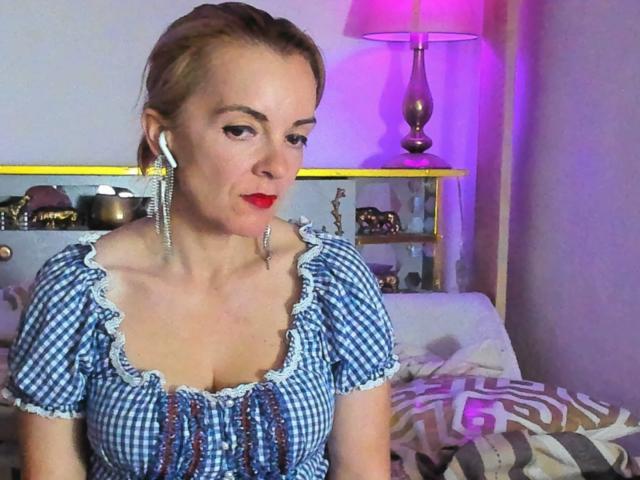 KateRouge with red lipstick, blue checkered dress, ruffled collar, and long silver earrings, showing her tits, in a bedroom with purple lamp light.