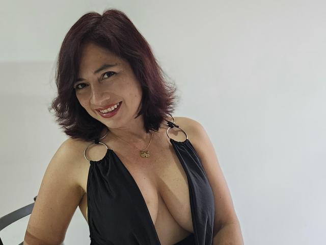 AlessiaRed has medium-length dark red hair, smiling, wearing a low-cut black dress with metal rings, showing her big tits, and a gold necklace with a small pendant.