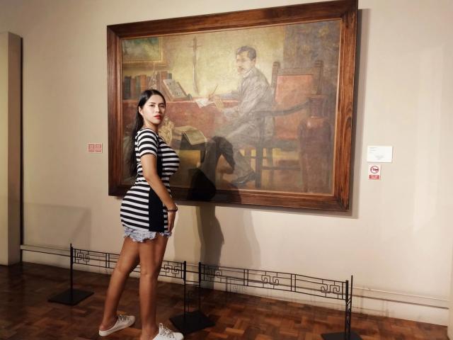 Sensualbunny-hot with striped shirt and denim shorts, standing in front of a large painting of a man with a book and chair, showing her ass and white sneakers.