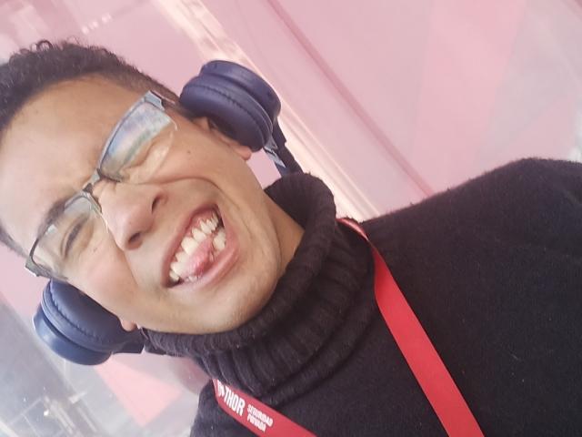 MartinxHt with glasses, grinning with teeth showing, wearing black turtleneck, blue headphones, red lanyard, and short curly hair. MartinxHt's face is slightly squished in a happy expression.