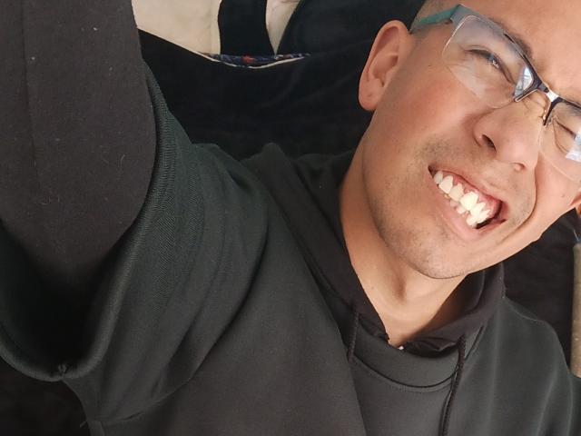 MartinxHt with glasses, black hoodie, and a big smile showing teeth, taking a selfie with his right arm raised. His skin is light brown and he's looking slightly to the side.