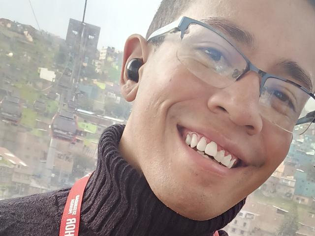 MartinxHt has a happy face, wearing glasses and a black turtleneck, with a red strap, and a black earbud, smiling with white teeth, short brown hair.
