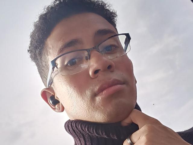 MartinxHt has curly black hair, wears glasses, and a black turtleneck, with a ring on his finger, and black earbuds. His skin is light brown, and he's looking up.