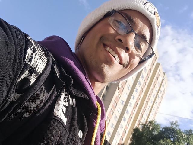 MartinxHt, smiling with glasses, white beanie, black jacket with white graffiti, purple hoodie, blue sky, and tall building in background. MartinxHt's ass is not visible.
