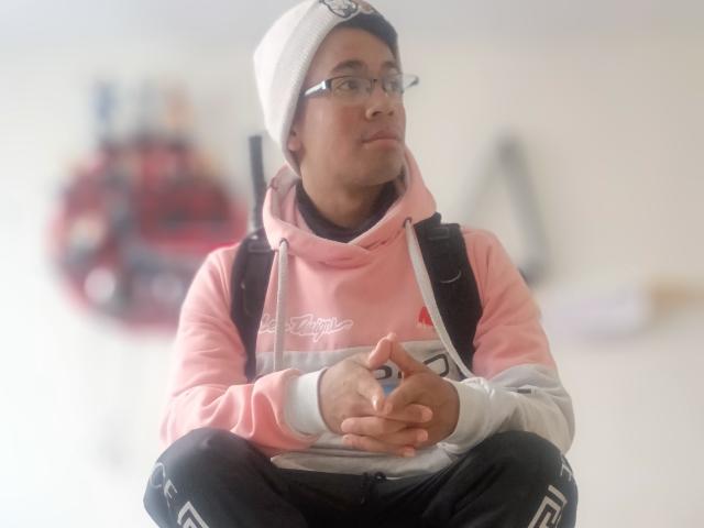 MartinxHt with glasses, white beanie, pink and gray hoodie, black pants, sitting cross-legged, hands together, backpack, looking up, blurry background, soft light, casual style.