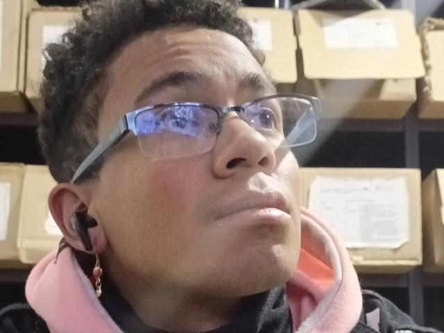 MartinxHt has curly black hair, wears blue glasses, and has a serious expression with slightly parted lips. He's in a pink hoodie with black earbuds.