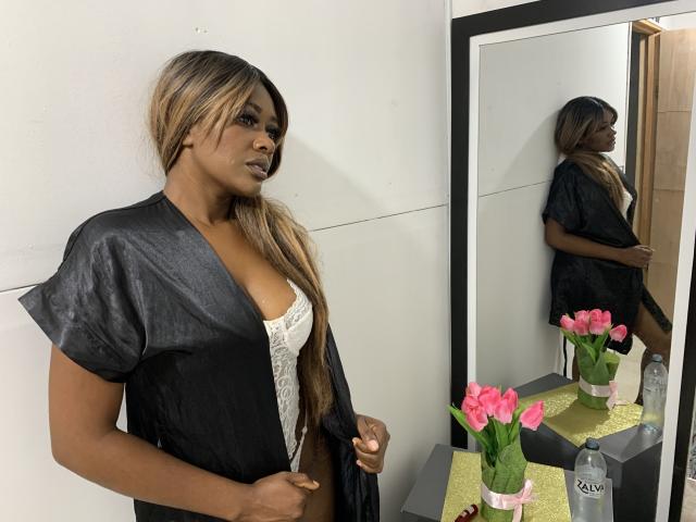 AstridDaviz with dark skin, long blonde hair, black satin robe, white lace bra, standing in front of mirror, reflection showing her ass, pink tulips on table, water bottle.