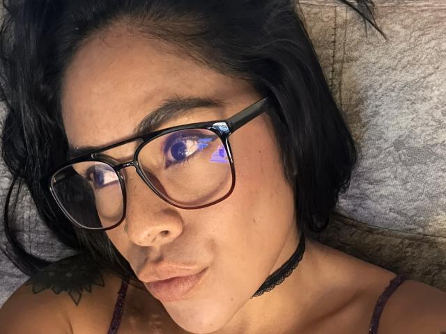 StellaBeckker with black glasses, purple eye reflection, choker, dark hair, tattoo on left shoulder, pouty lips, and a slightly flushed face. Boobs visible in purple strap top.