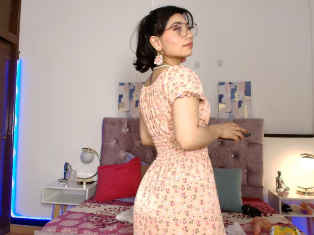 SeleneLeon with glasses, pink floral dress, pink earrings, looking over shoulder, standing in bedroom, showing her ass, short black hair, light skin, bed with pink and blue pillows.