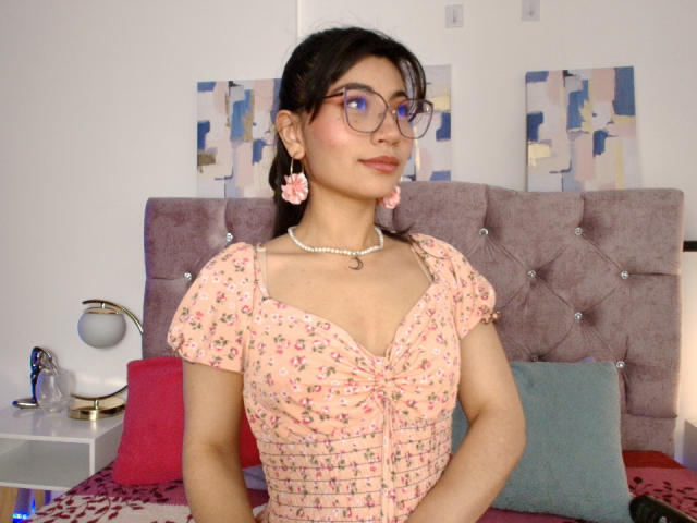 SeleneLeon with dark hair, glasses, pink floral dress, pink earrings, pearl necklace, small tits, sitting on bed, pink pillow, purple headboard, blue and white wall art.