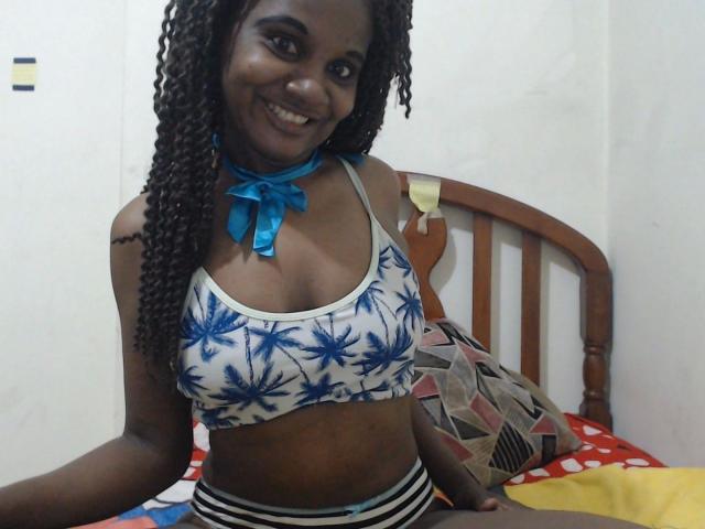 Kathrina87-hot, dark-skinned with curly hair, smiling, wearing a blue-patterned sports bra and striped underwear, blue ribbon choker, sitting on a bed with a patterned pillow.