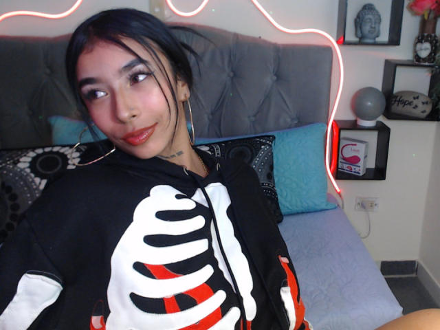 MaddyParker with dark hair, wearing a black skeleton hoodie, sitting on a bed with red LED lights, slightly smiling, showing a small tattoo on her neck.