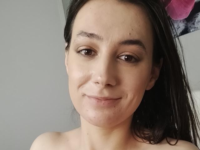 JuliaHuntis with light skin, brown eyes, and straight black hair, smiling softly at the camera, showing subtle wrinkles around her mouth. She's topless, with bare shoulders visible.