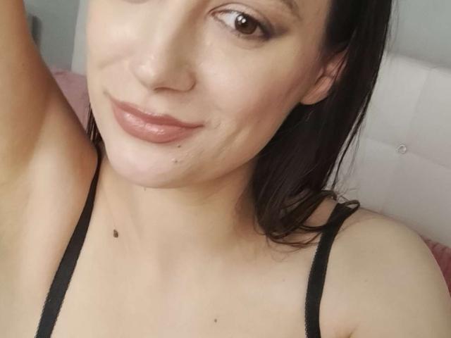 JuliaHuntis has light skin, brown eyes, and a small black mole below her chin. She's wearing a black strap top with her arm raised, smiling slightly.