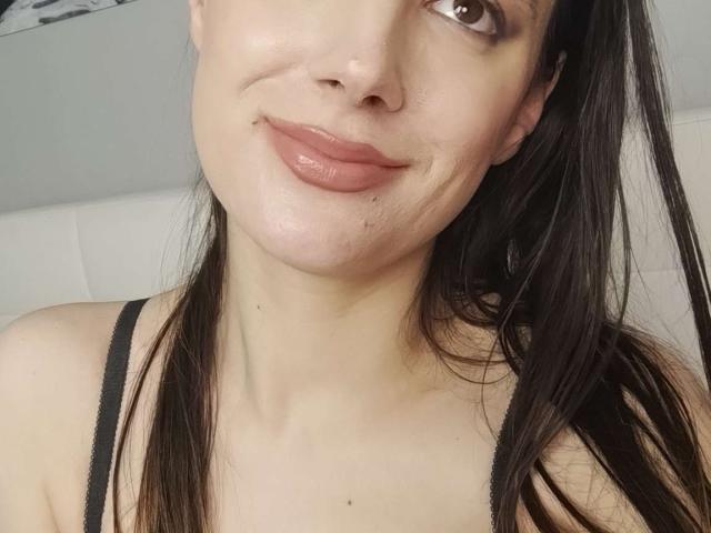 JuliaHuntis tem pele clara, olhos castanhos e lábios rosados e cheios, com um leve sorriso. Ela está usando uma alça preta, e seu cabelo castanho escuro está solto..