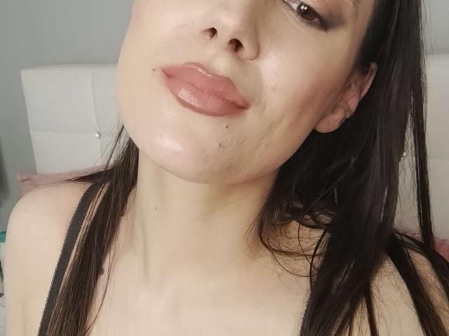 JuliaHuntis with glossy pink lips, slightly shiny skin, small dark moles, dark brown hair, wearing a black strap, head tilted. Boobs not visible, focus on face.
