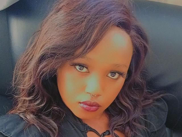 Wetnorah has dark brown wavy hair, a nose ring, and red lipstick, with long eyelashes and brown eyes, wearing a black jacket, looking straight at the camera.