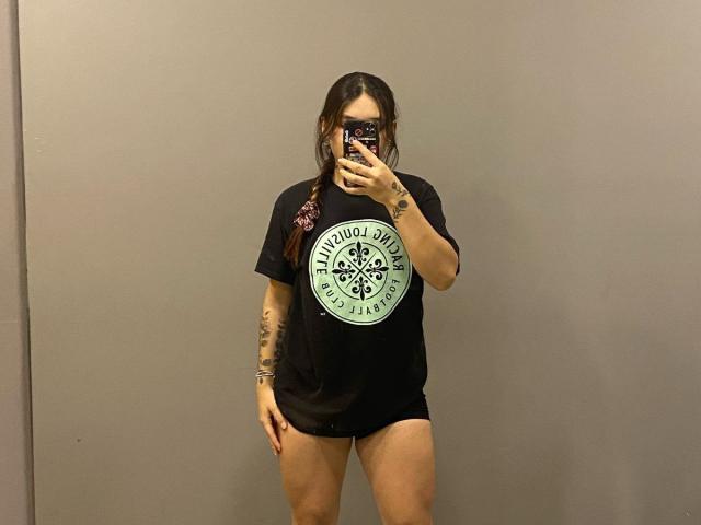 SophiaMarx with long brown hair in a braid, wearing a black "Knot Dead" tee, black shorts, mask, tattoos on arm, holding phone, taking mirror selfie, showing her ass.