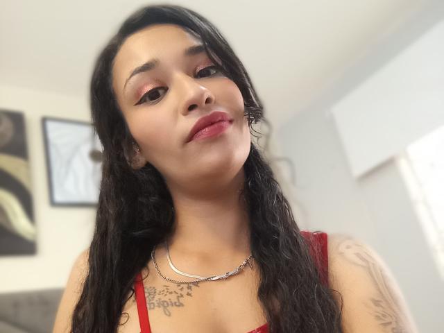 Zoey Moretti mit welligem, schwarzem Haar, rotem Lippenstift und einem roten Kleid, die Tattoos auf ihrer Brust zeigt, eine silberne Halskette trägt und mit einem leichten Lächeln in die Kamera schaut..