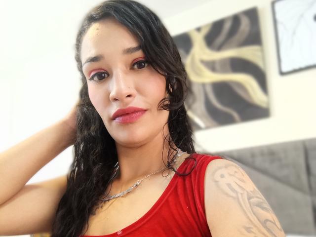 Zoey Moretti, com cabelo preto molhado, batom vermelho e blusa vermelha, exibindo tatuagens no braço direito, usando um colar de prata, com a mão no cabelo, olhando para a câmera..