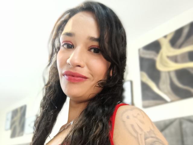 Zoey Moretti hat lockiges, schwarzes Haar, roten Lippenstift, ein Tattoo an ihrer rechten Schulter und trägt ein rotes Oberteil. Sie lächelt und hat einen Nasenring..