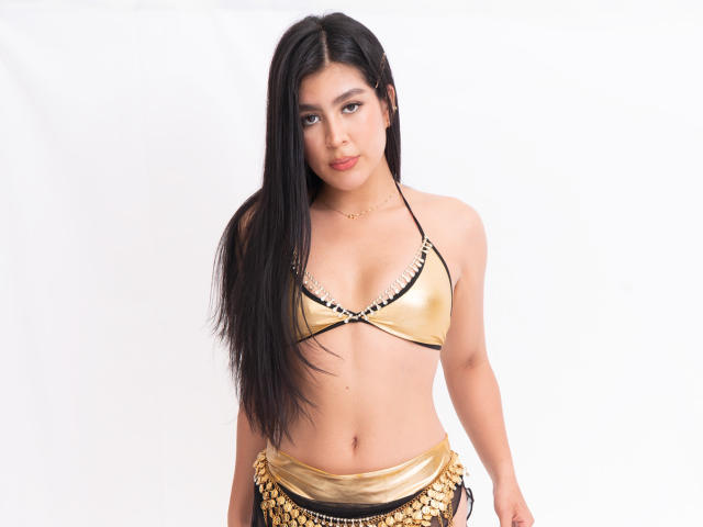 SamanthaEva69 with long black hair, gold bikini top, and gold chain belt, standing, showing her tits, and a slight smile, against a white background.
