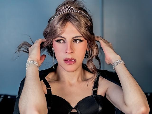 LuAngeli mit hochgezogenen Lippen, Händen im braunen Haar, trägt einen schwarzen BH, eine Tiara und ein glitzerndes Armband und blickt in die Kamera. Die Brüste sind sichtbar, sie trägt pinken Lippenstift und hat braune Augen..