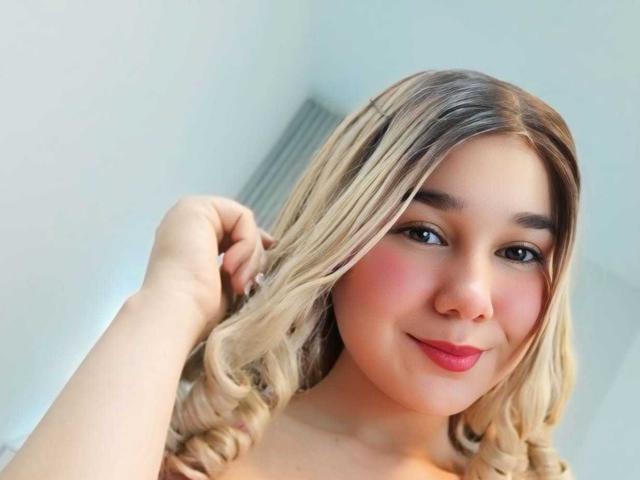 IvanaStaysi mit blonden, gewellten Haaren, rosiger Rouge, rotem Lippenstift und einem süßen Lächeln, die ihre Haare berührt. Sie ist oben ohne, wobei ihre Brüste teilweise sichtbar sind. Weiches, natürliches Licht..