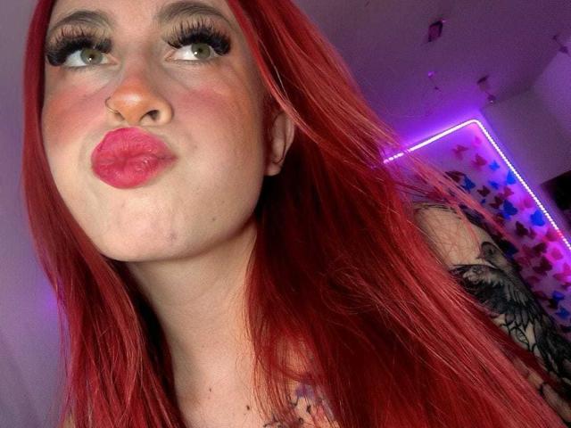 SammyViolet with red hair, pouty pink lips, green eyes, and long eyelashes, making a duck face, showing a tattoo on her right shoulder, in a selfie.