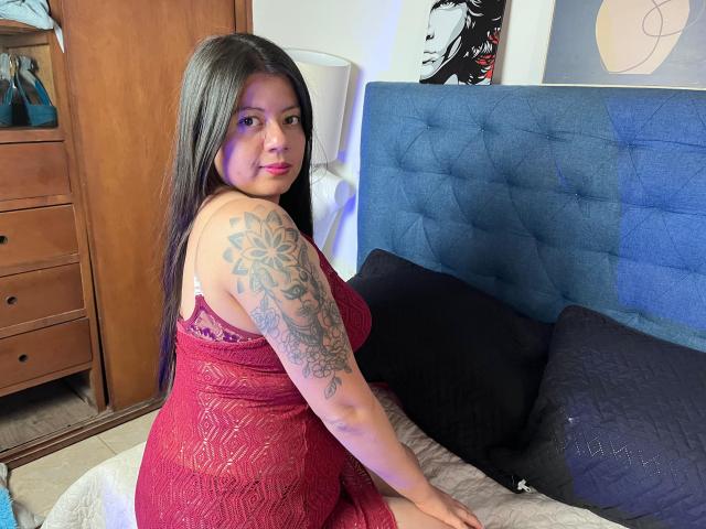 Lily Parker, avec de longs cheveux bruns, une robe rouge en dentelle, montrant un tatouage sur le bras droit, assise sur un lit, regardant par-dessus son épaule, avec une tête de lit bleue, des oreillers noirs, une commode en bois et une lampe blanche..