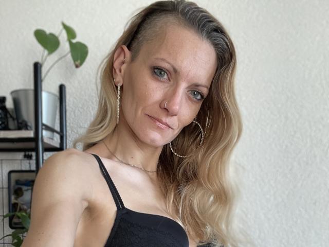 DaneeSoHot with side-shaved blonde hair, hoop earrings, black lace top, subtle nose ring, and a confident smirk. Her tits are small, and she's looking at the camera.
