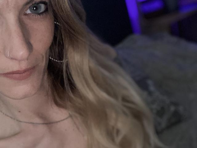 DaneeSoHot's face with blue eyes, nose ring, and blonde hair, wearing thin earrings and necklace, in a dimly lit room. Boobs not visible.