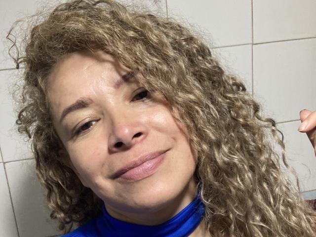 DalilaSuperHot with curly blonde hair, light skin, and a slight smile, wearing a blue top, showing a cute nose and pinkish lips, holding a finger to the right.