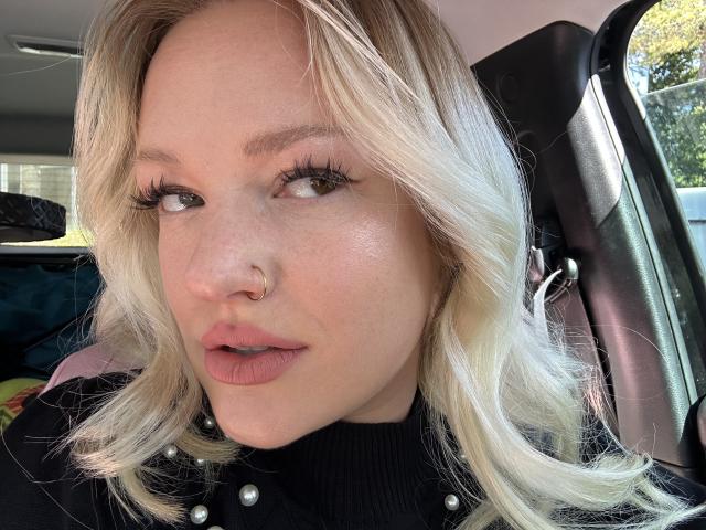 Cherry7up has blonde wavy hair, a nose ring, and long eyelashes, wearing a black shirt with pearl buttons, looking to the side with slightly parted pink lips.