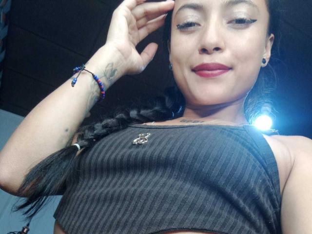 MaiteX porte un débardeur noir à rayures, qui met en valeur sa petite poitrine, ses cheveux noirs tressés, son rouge à lèvres rouge, un tatouage au poignet et un bracelet de perles, et elle sourit à l'appareil photo..