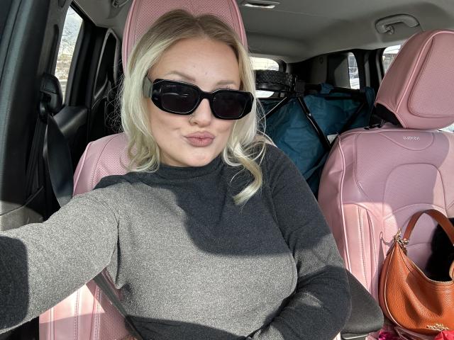Cherry7up with blonde hair, black sunglasses, and a grey sweater, sitting in a pink car seat, showing pouty lips, with a brown bag and blue luggage in the background.