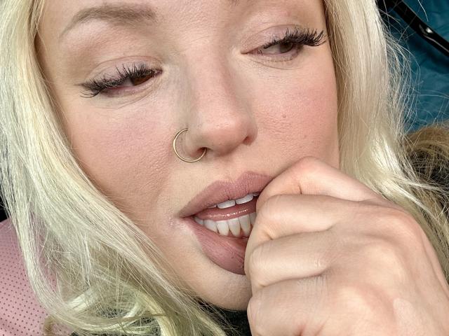 Cherry7up with blonde hair, nose ring, and pink lips, biting her finger, showing white teeth, and wearing long eyelashes. Her hand is near her mouth.