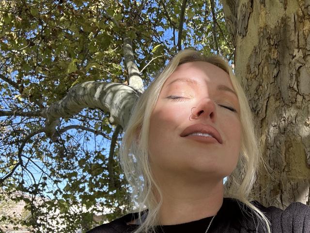Cherry7up with eyes closed, nose pierced, blonde hair, black top, sunlit face, relaxed expression, standing by a textured tree, blue sky, green leaves in the background.