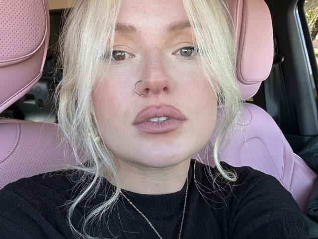 Cherry7up with blonde hair, nose ring, and pink leather car seat. She's wearing a black shirt, looking straight at the camera with slightly parted pink lips.