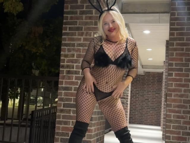 Cherry7up with blonde hair, red lipstick, black fishnet bodysuit, black bunny ears, black bra, black panties, black thigh-high boots, standing in brick doorway at night.