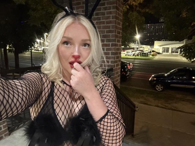 Cherry7up with blonde hair, fishnet top, black bunny ears, and red lipstick, biting her finger, showing her tits, outside at night, in a selfie.