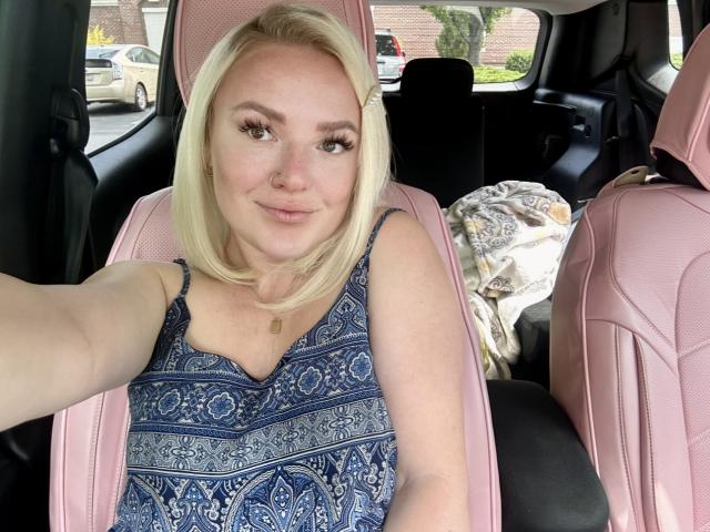Cherry7up, blonde, in pink car seat, wearing blue patterned dress, smiling, holding phone for selfie, pink car interior, patterned blanket in background, visible nose ring.