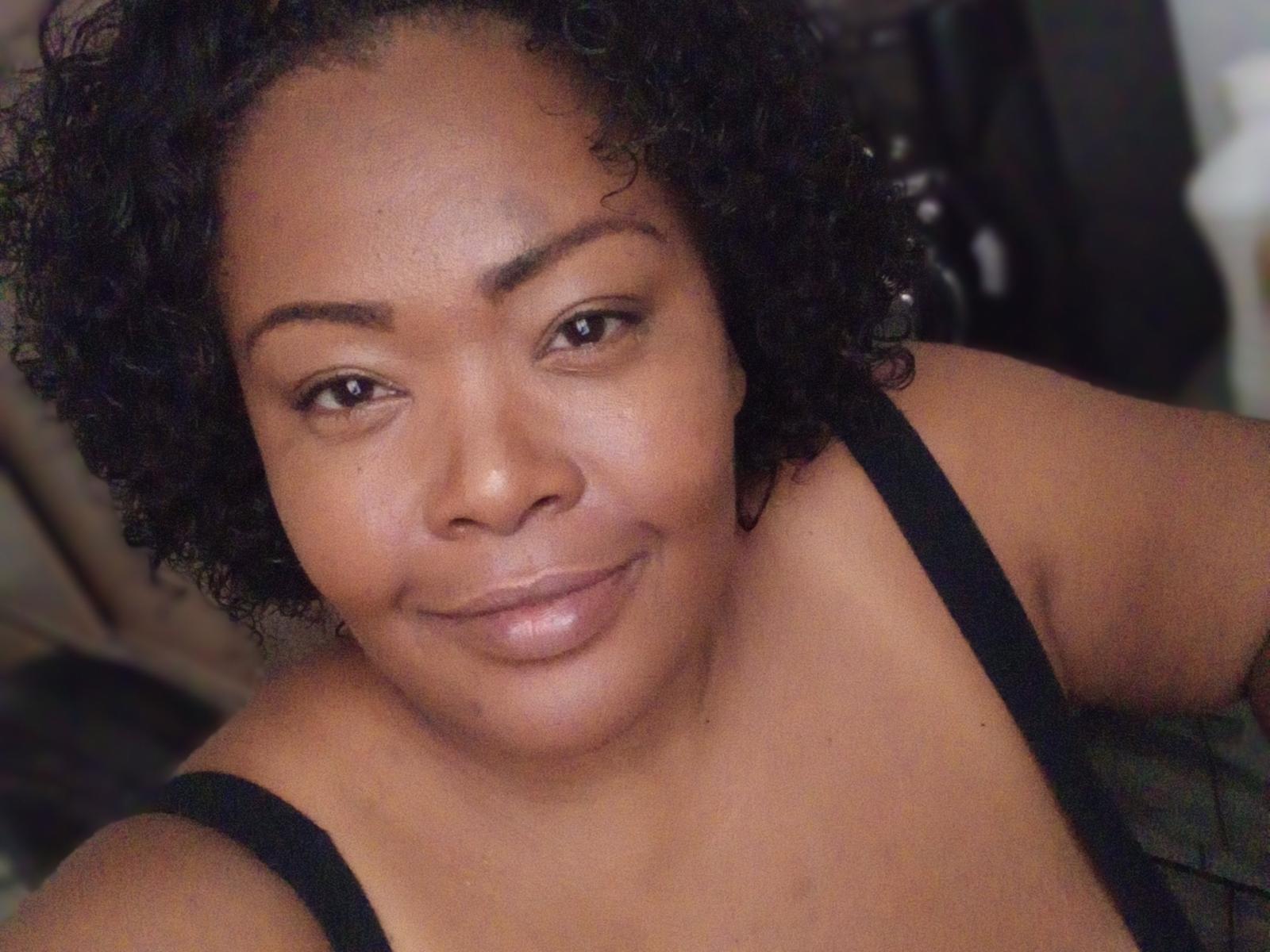 Lilith69z has curly black hair, dark brown skin, and is smiling softly at the camera, wearing a black strap top, showing her small, round tits and a cute face.