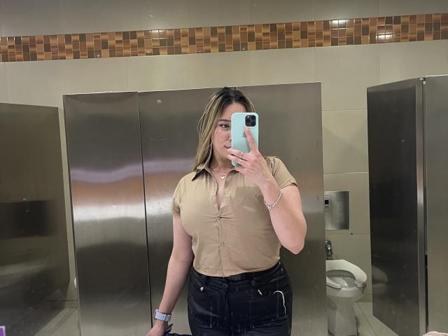 JennyLawry, con un seno di media grandezza, indossa una camicia beige con bottoni e jeans neri, e sta scattando un selfie allo specchio in un bagno con pareti divisorie in acciaio inossidabile, tenendo in mano un telefono blu..