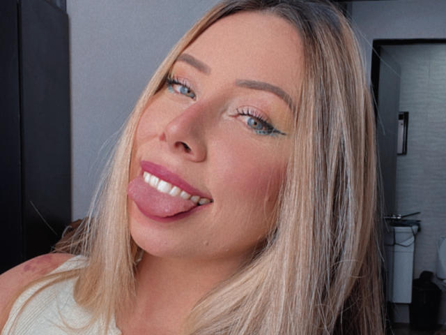 JennyLawry's face, blonde hair, blue eyes, pink lipstick, tongue out, smiling, white tank top, light makeup, slightly flushed cheeks, soft focus, casual selfie.
