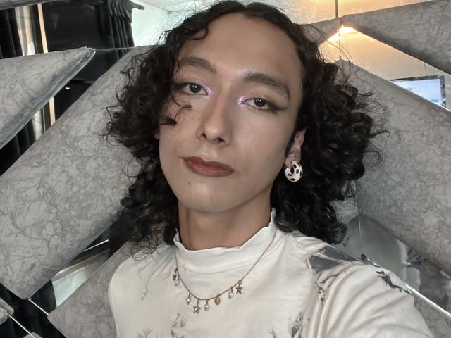 MissJami69 has curly black hair, light brown skin, purple eyeshadow, a small bandage on her cheek, and wears a white top with a star necklace, and a skull earring.